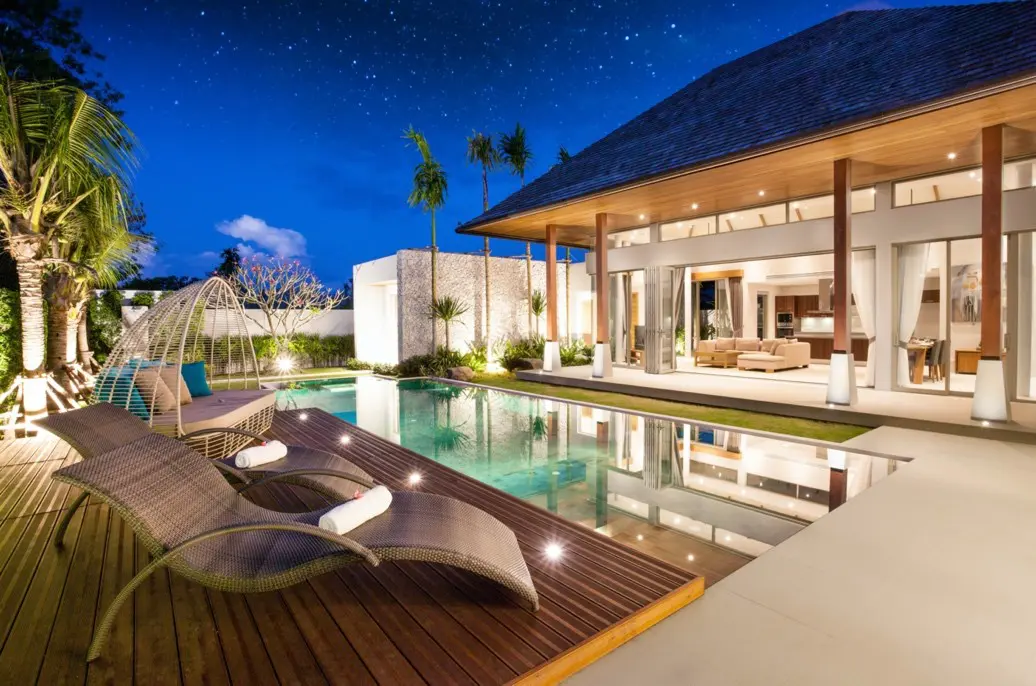 A pool with lounge chairs and a house in the background.